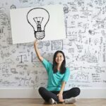 Une jeune femme tient un dessin d'ampoule contre un mur de brainstorming créatif.